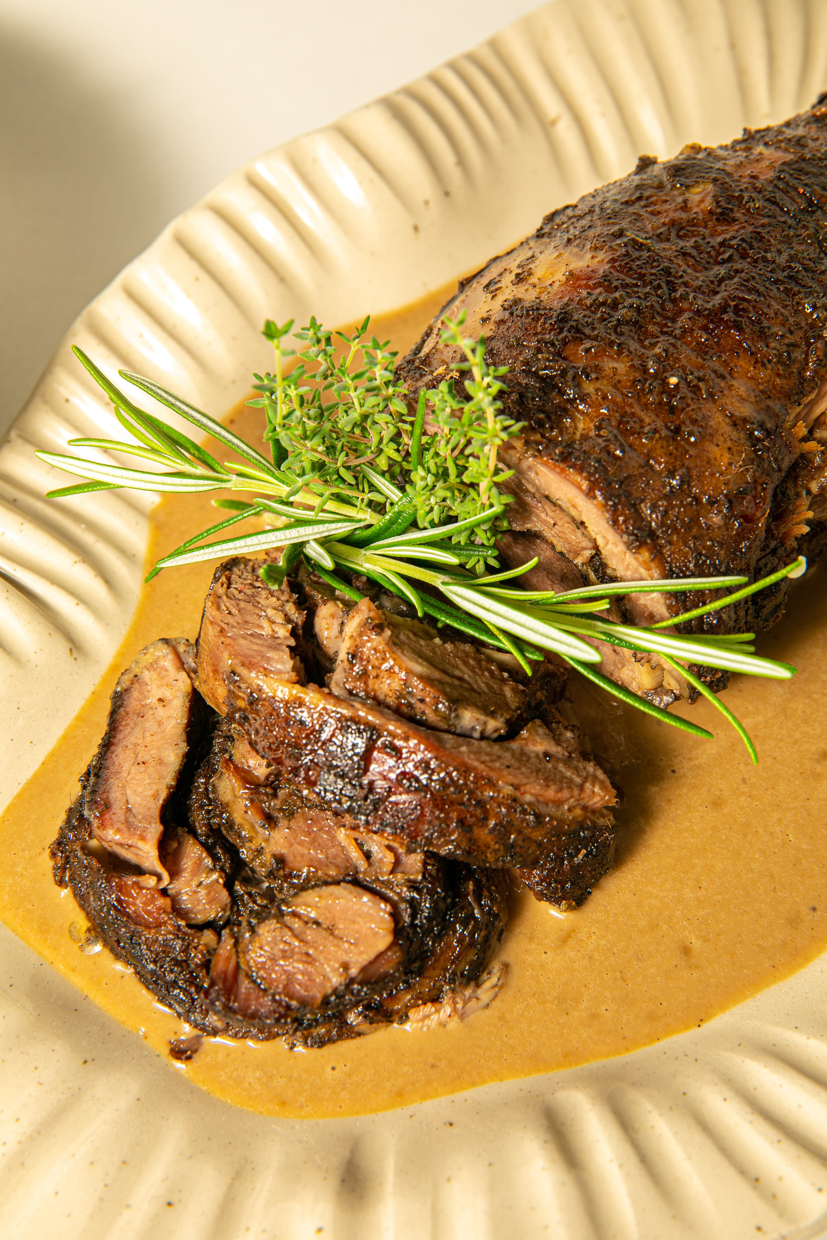A whole spice-crusted roast lamb shoulder on a fluted cream platter, garnished with rosemary and thyme in a pool of golden jus.
