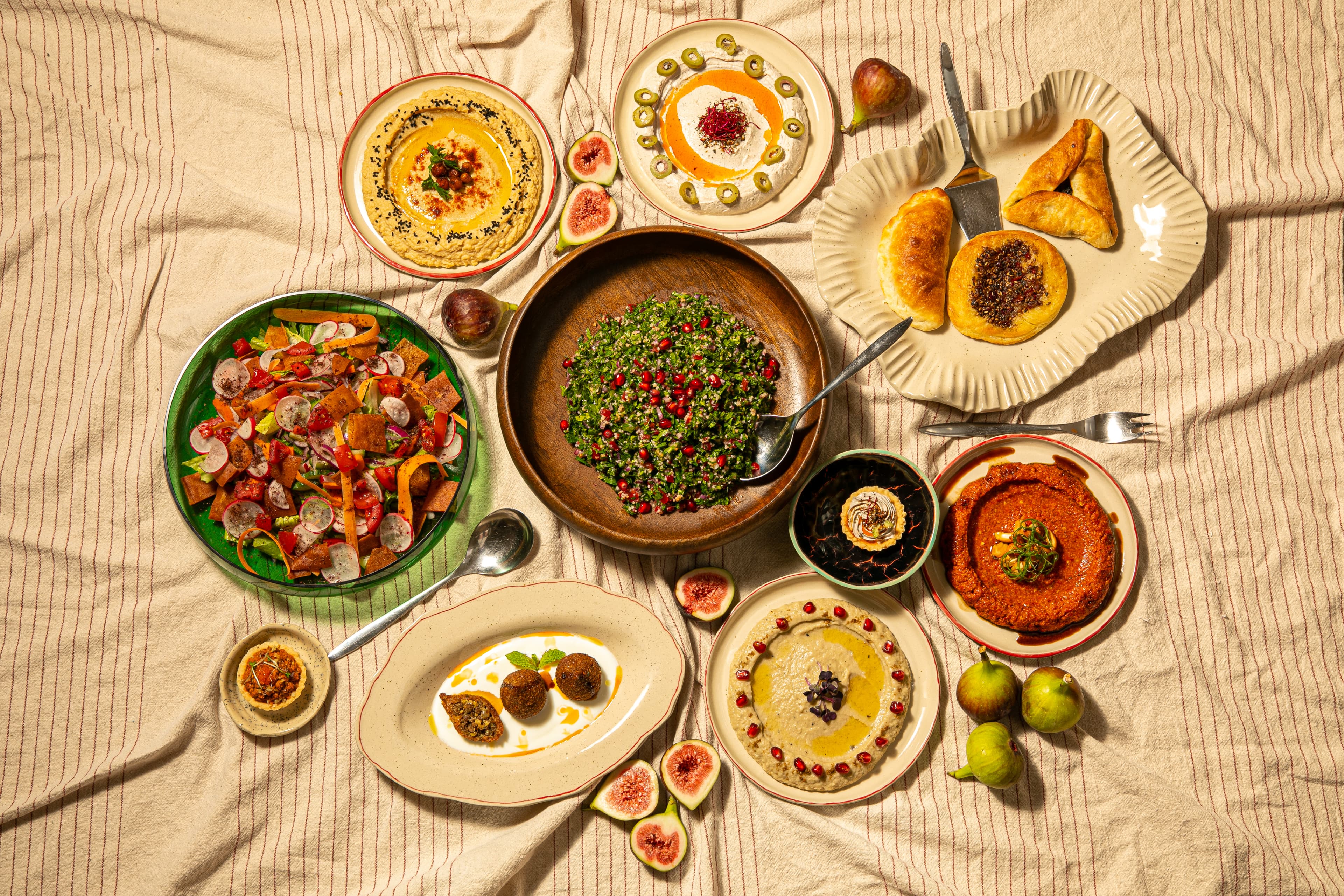 An overhead spread of Levantine mezze — hummus, tabbouleh, muhammara, fatteh, fresh salads, savoury pastries and figs laid across a striped linen tablecloth.