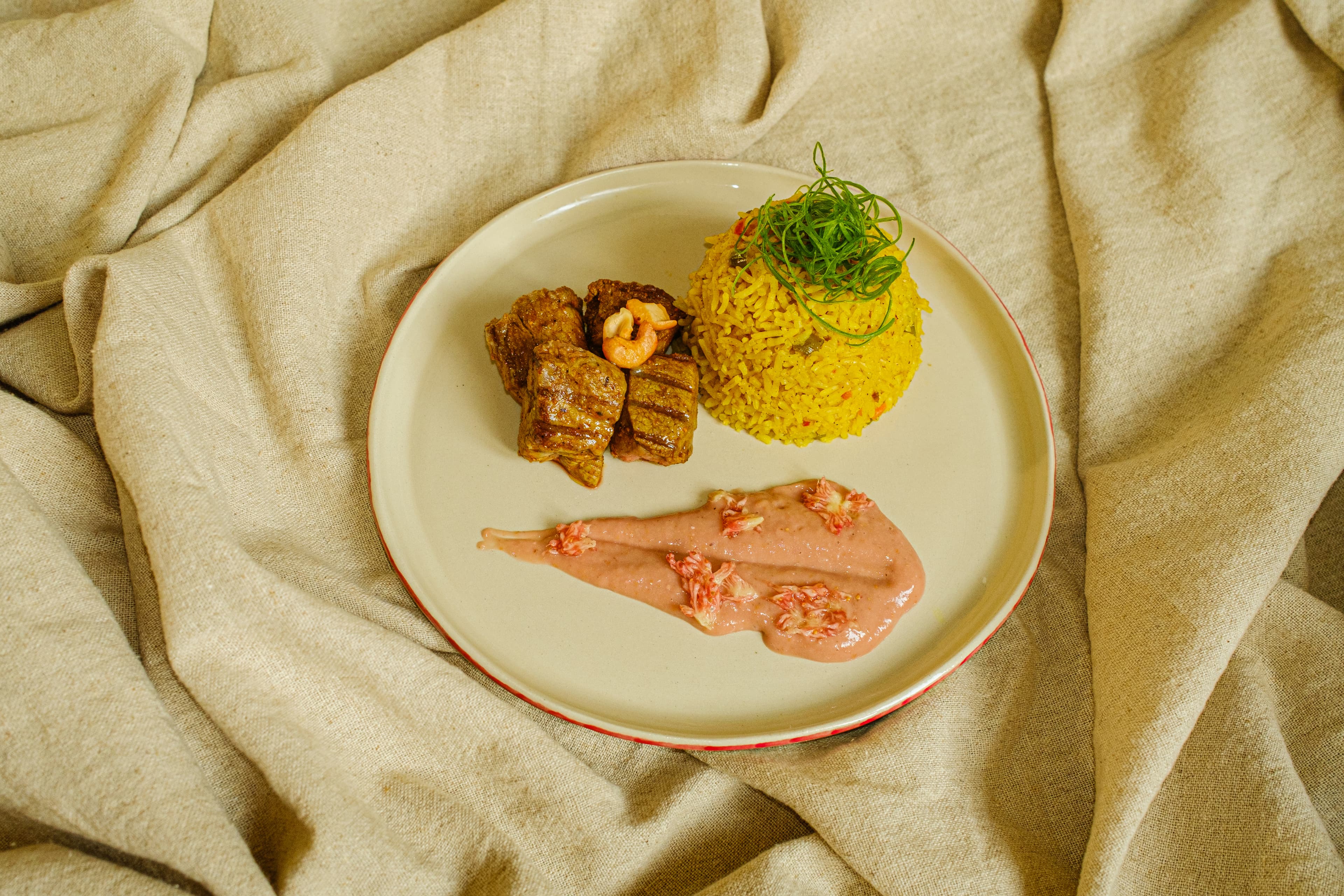 A plated main course on a red-rimmed cream plate — seared beef medallions with a prawn, a mound of saffron rice topped with crispy leek, and a smear of pink sauce — set on a linen tablecloth.