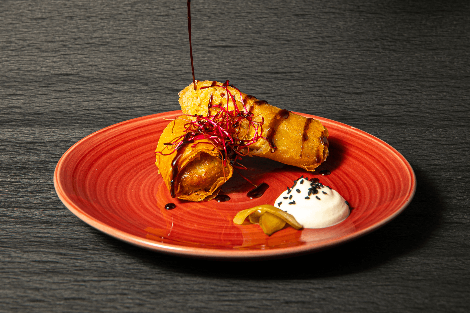 A crisp golden pastry roll on a rustic red plate, drizzled with pomegranate molasses and served with labneh and pickled chilli.