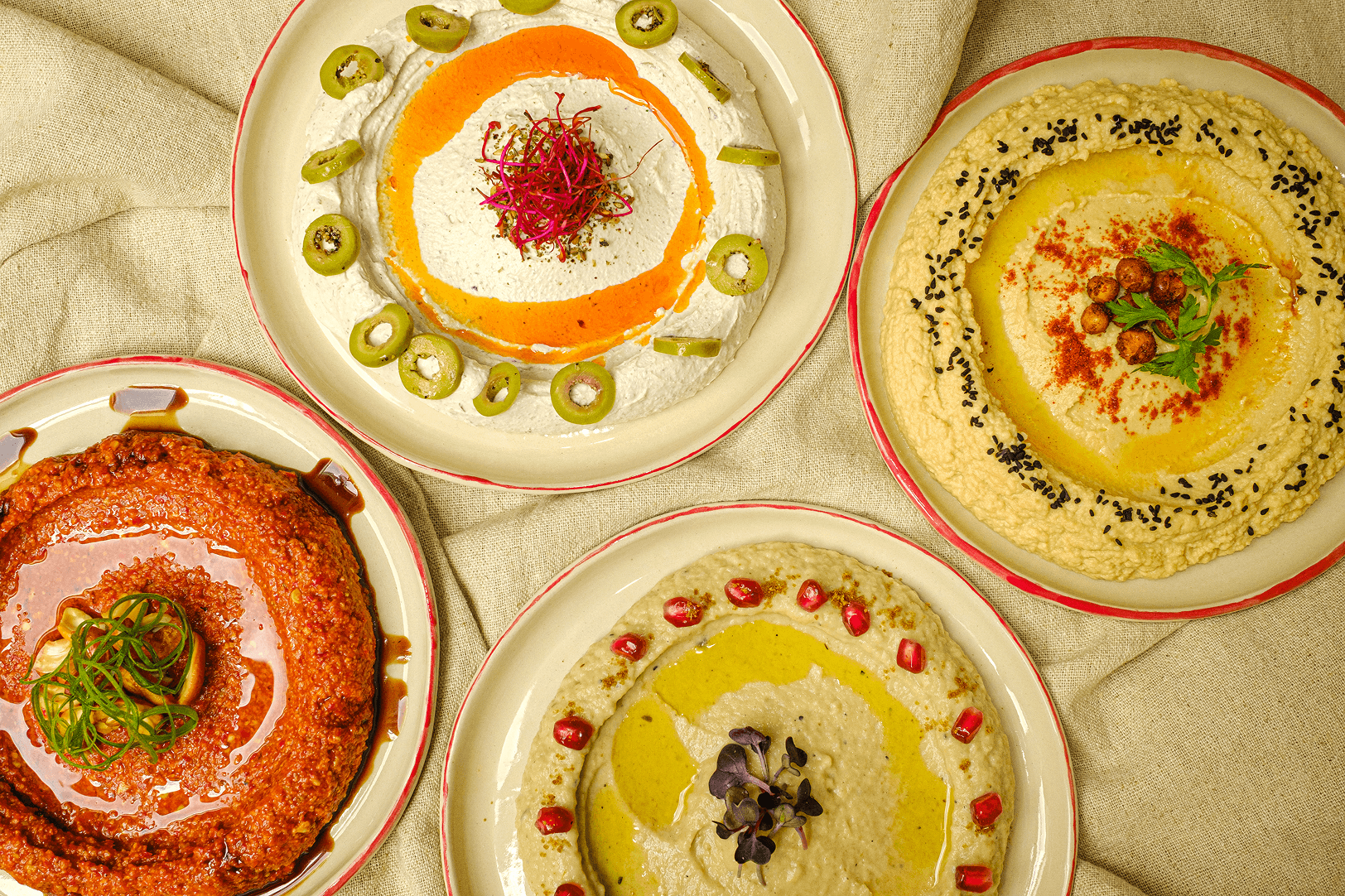 An overhead spread of four mezze dips — hummus, muhammara and labneh — garnished with olives, pomegranate and black sesame on a linen cloth.