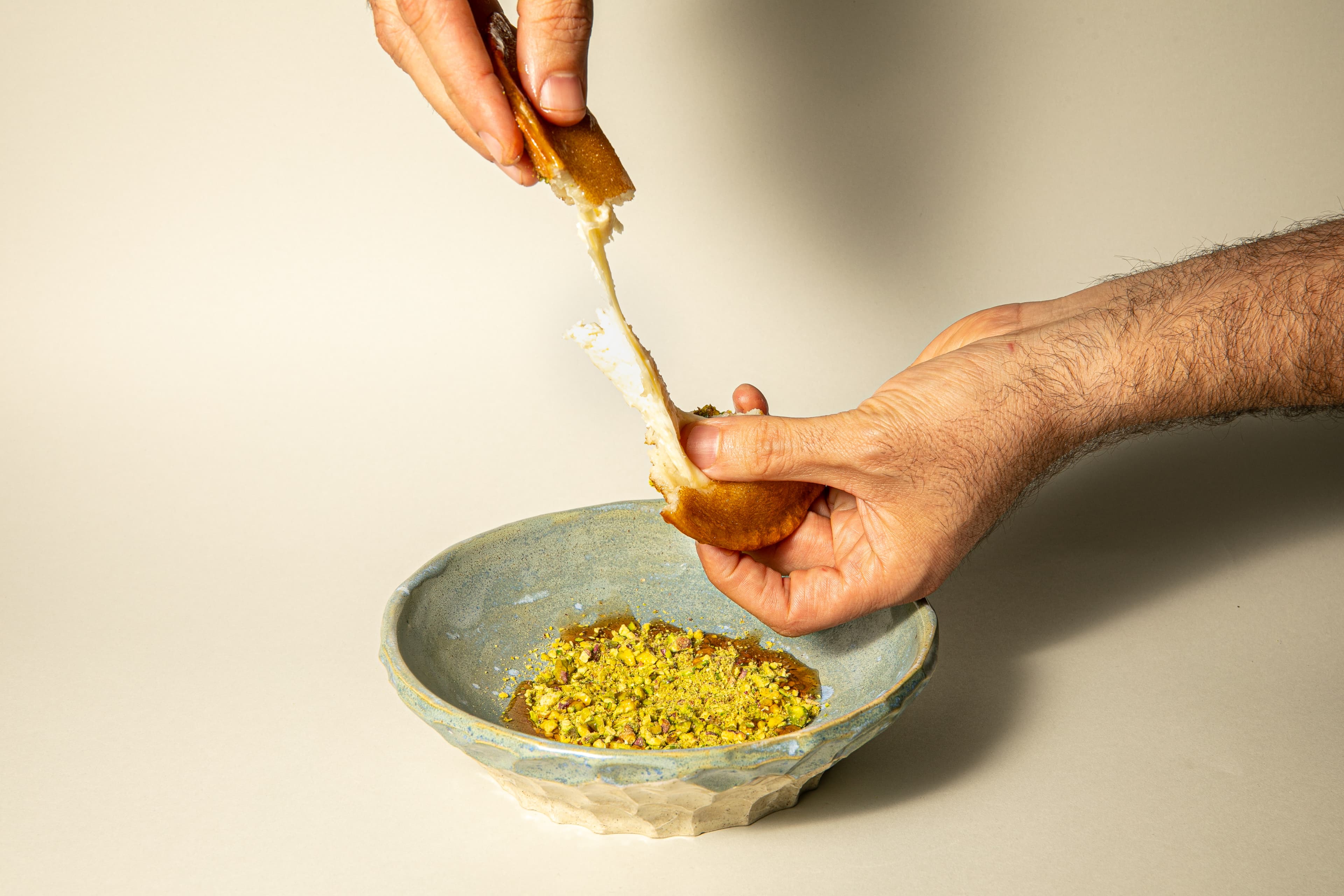Two hands pulling apart a warm knafeh over a stone bowl of crushed pistachios.